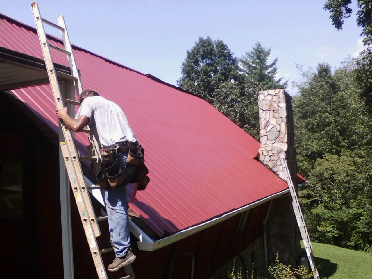 Roofer on a ladder beside a red metal roof and stone chimney in Yale